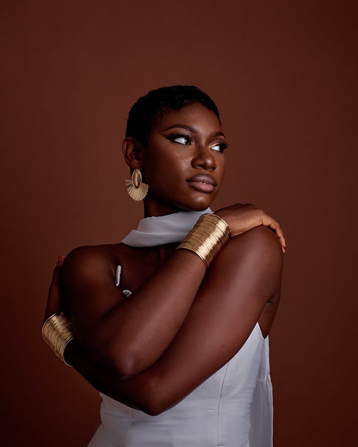 Stylish woman in white dress with gold jewelry against brown background.
